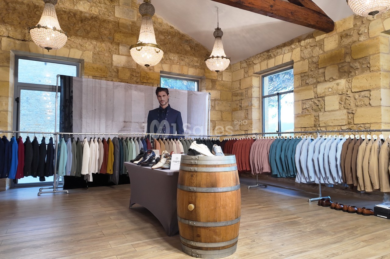 Salle avec des vêtements suspendus sur des portants, chaussures exposées sur une table et un tonneau, murs en pierre…