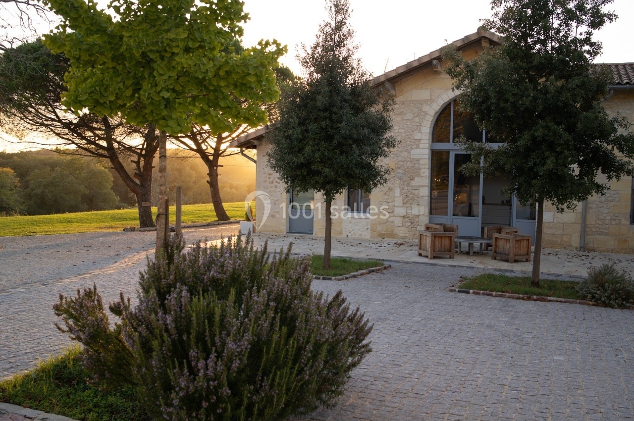 Façade d'un bâtiment en pierre avec grandes fenêtres, entouré d'arbres et d'un espace pavé au coucher du soleil.