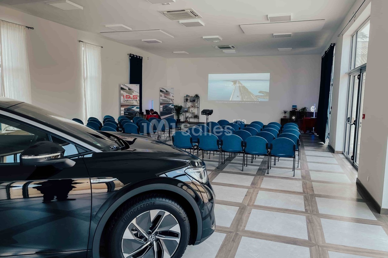Salle de conférence lumineuse avec des rangées de chaises bleues, un écran de projection et une voiture au premier plan.