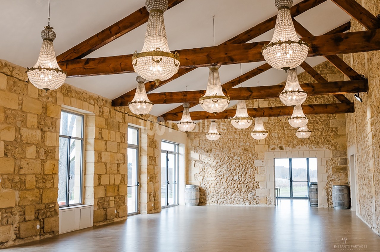 Salle spacieuse avec murs en pierre, poutres apparentes et lustres suspendus, éclairée par de grandes fenêtres.