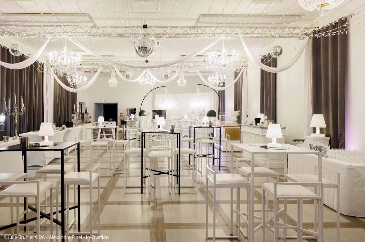 Salle de réception décorée avec des tables hautes, des guirlandes lumineuses et des lustres suspendus.