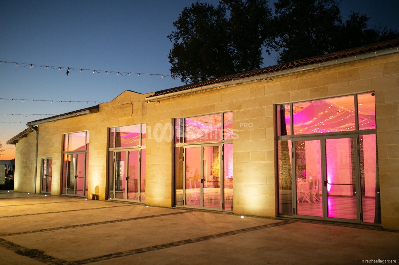 Façade d'un bâtiment en pierre avec grandes baies vitrées illuminées par des lumières roses à la tombée de la nuit.