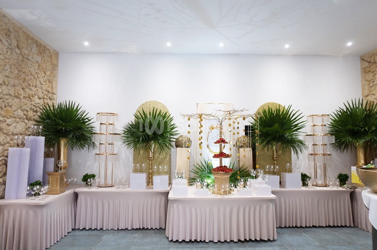 Table de buffet élégante décorée de palmiers, de dorures et de présentoirs à desserts dans une salle aux murs en pierre.