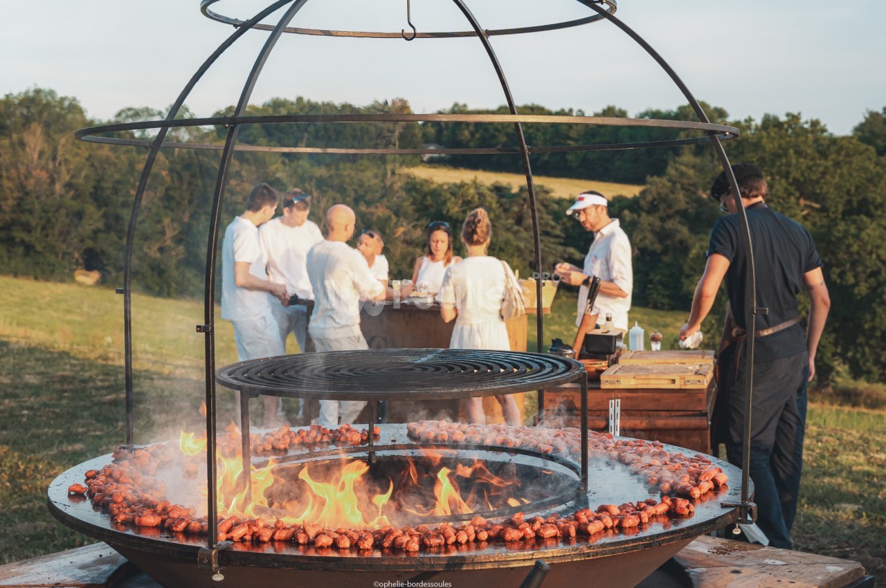 Des personnes discutent autour d'un barbecue circulaire en plein air, avec des saucisses grillant sur le feu.