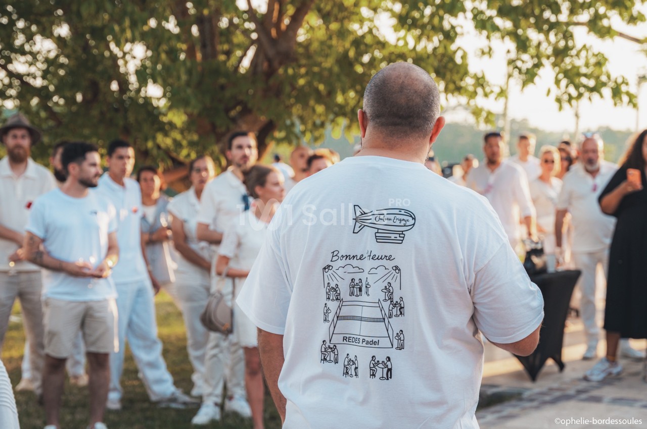 Un groupe de personnes en tenue blanche rassemblées en extérieur, un homme de dos portant un t-shirt illustré.