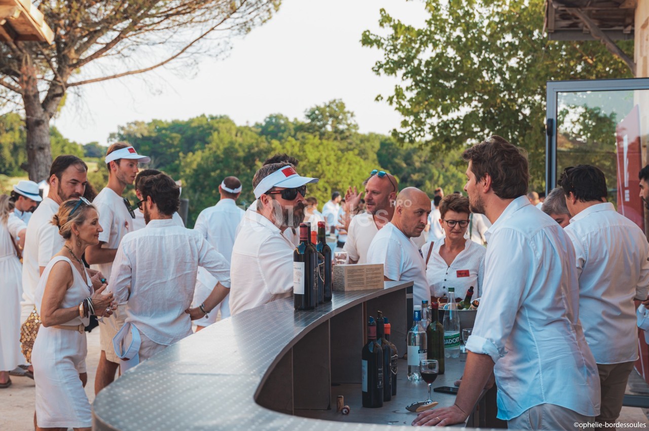 Des personnes en tenue blanche discutent et dégustent du vin autour d'un comptoir extérieur lors d'un événement.