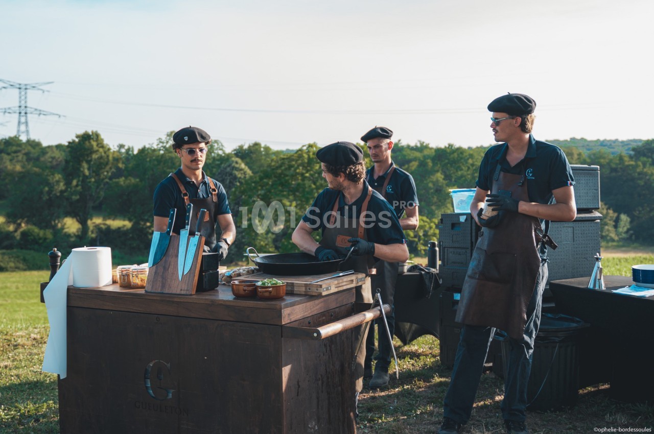 Quatre cuisiniers en tenue professionnelle préparent un repas en plein air devant un paysage verdoyant.