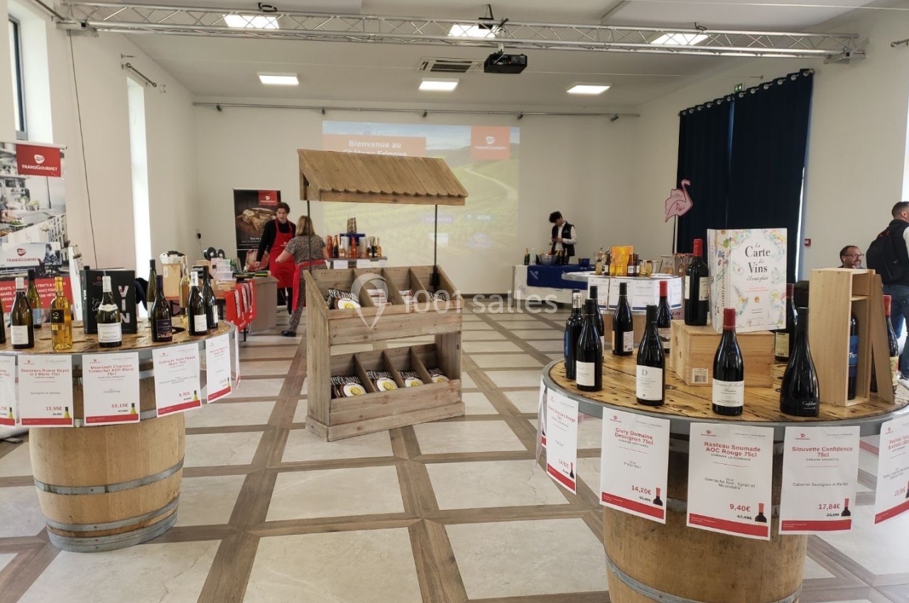 Salle d'exposition avec des stands de produits locaux, des bouteilles de vin sur des tonneaux et des étals de spécialités…