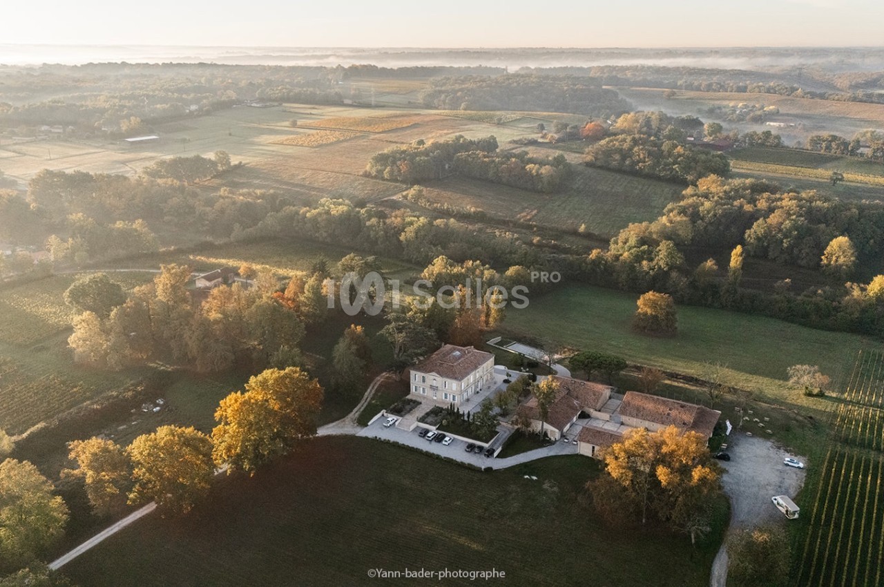 Vue aérienne d'un domaine entouré de champs, de vignes et d'arbres, baigné par une lumière douce au lever du soleil.