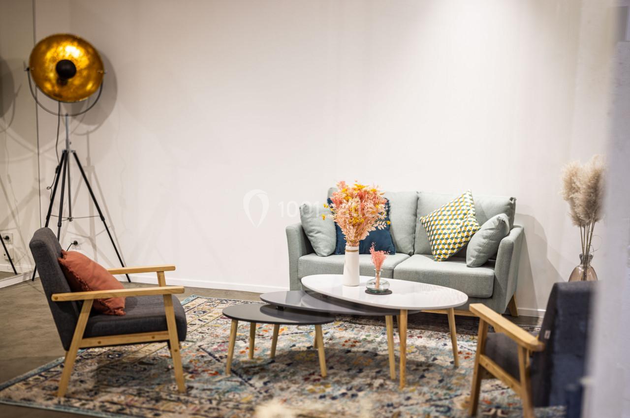 Salon moderne avec canapé vert, fauteuils gris, tables basses ovales et vase de fleurs colorées sur un tapis décoratif.