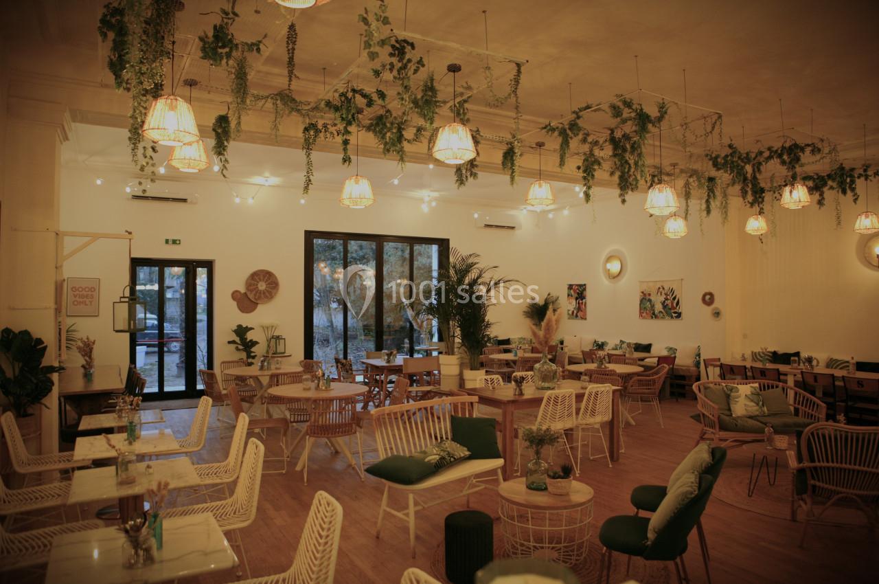 Salle de restaurant chaleureuse avec mobilier en rotin, plantes suspendues et lumière tamisée.
