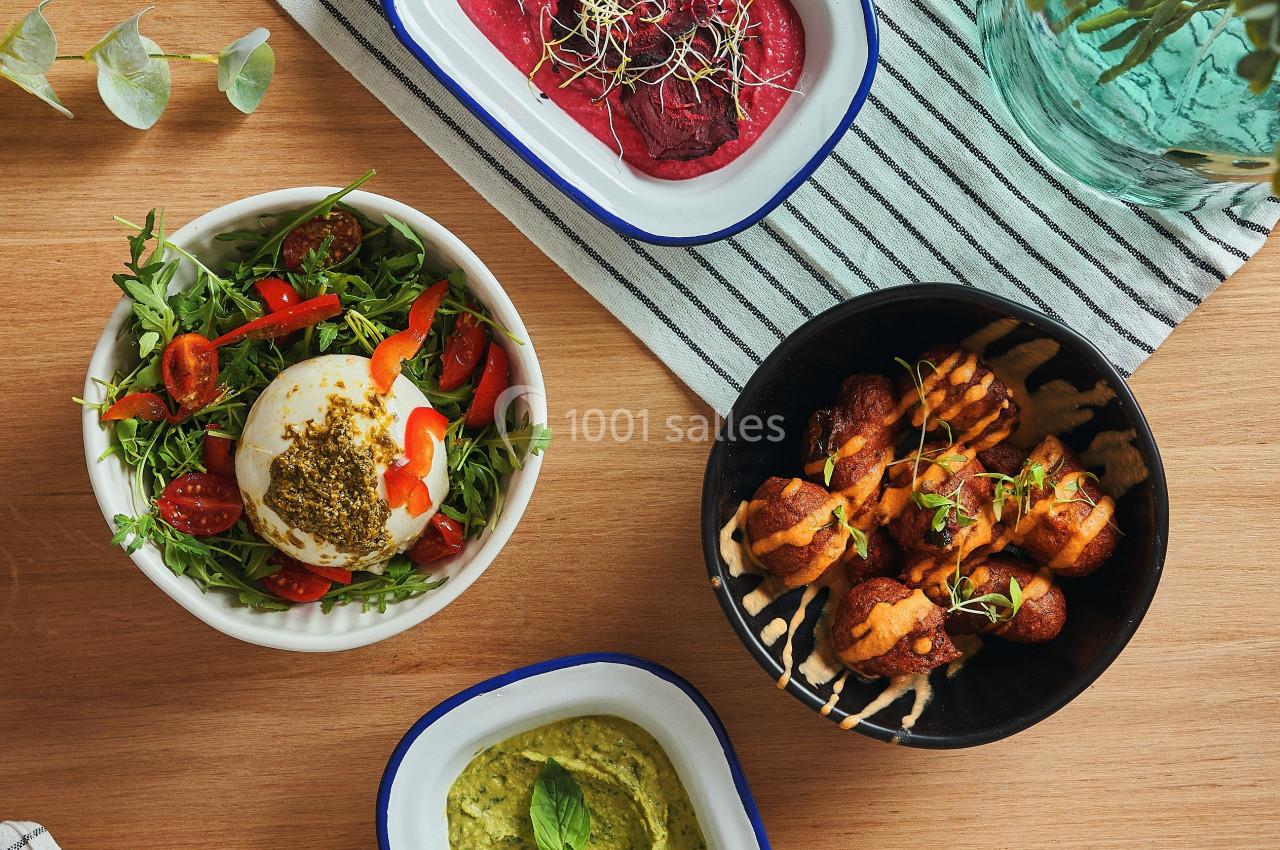 Quatre plats variés sur une table en bois : salade, houmous, brochettes et tartinade rouge, avec des couverts et une nappe.