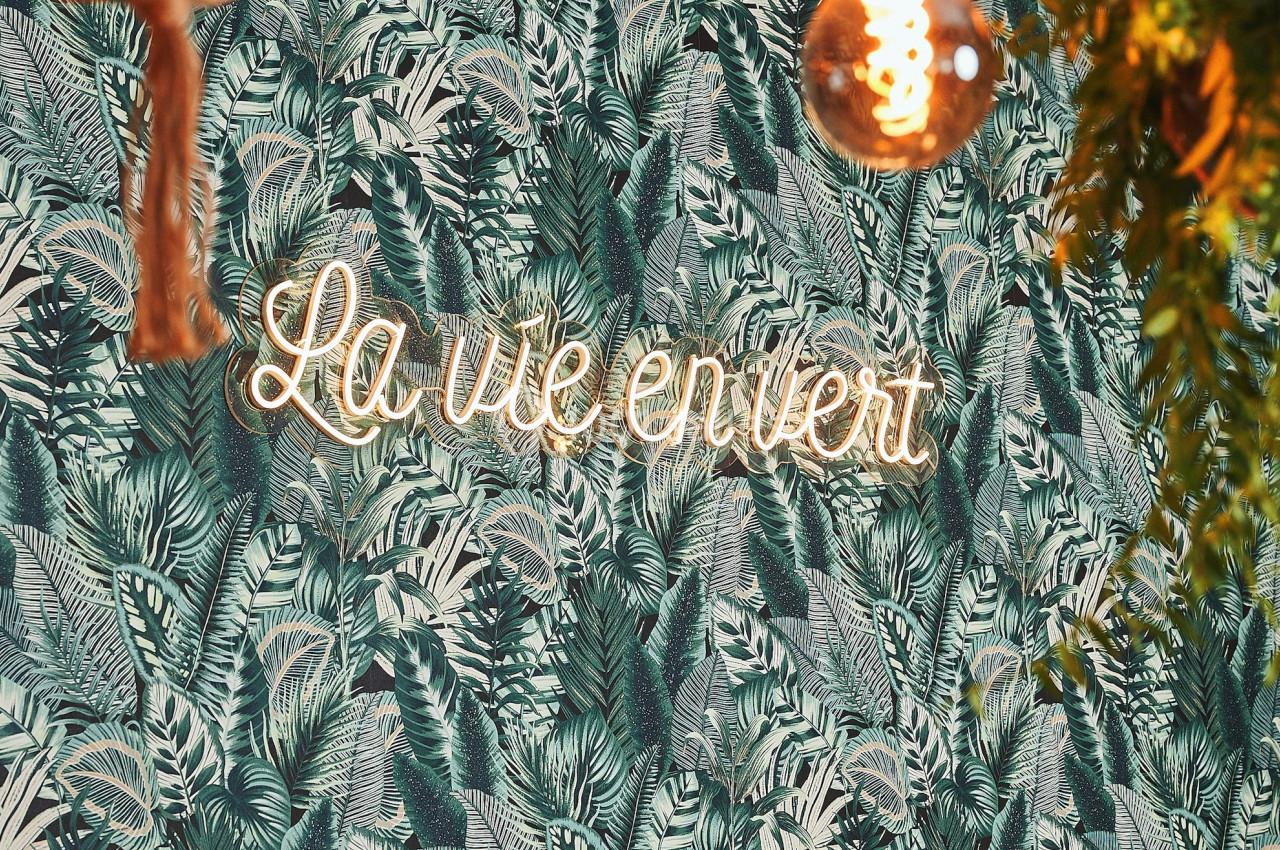 Mur décoré d'un papier peint à motif végétal, éclairé par une enseigne néon ’La vie en vert’ et des suspensions lumineuses.