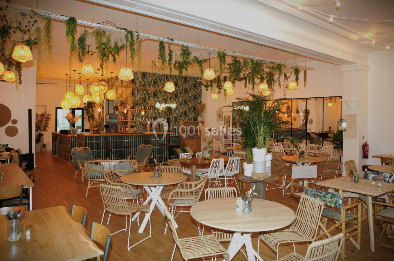 Salle de restaurant lumineuse avec mobilier en bois et rotin, décorée de plantes suspendues et lampes chaleureuses.