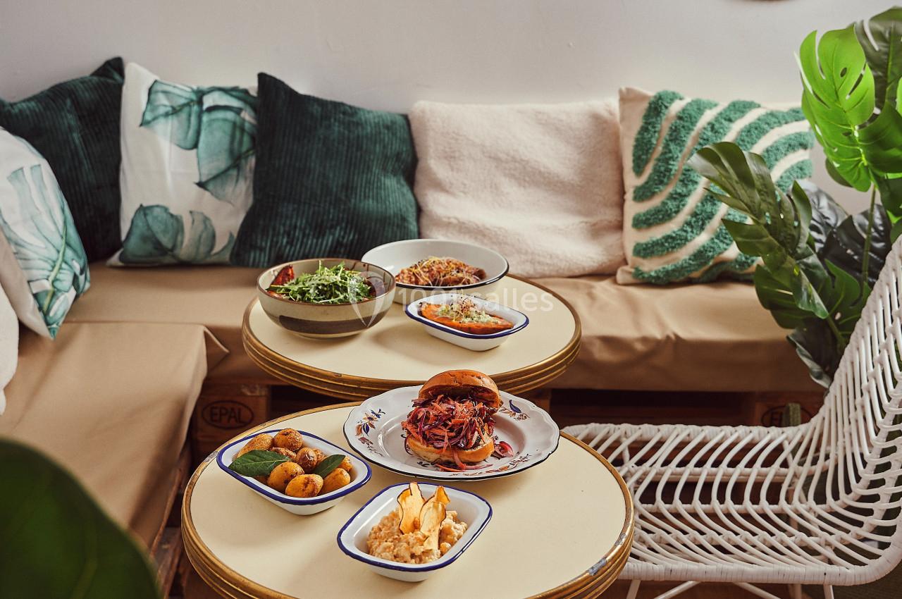 Assiettes de plats variés disposées sur des tables rondes dans un salon décoré de coussins et d'éléments naturels.
