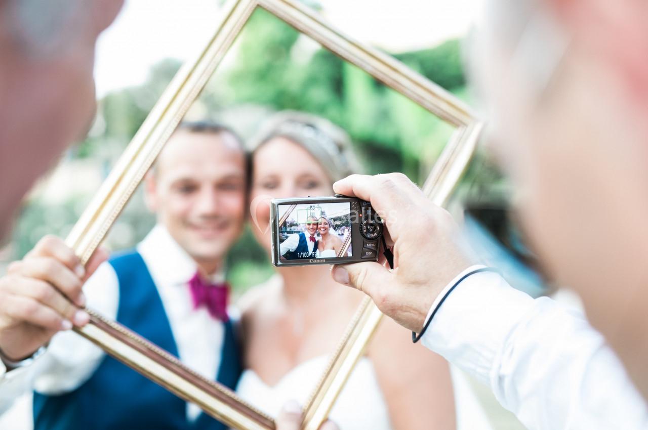 Un couple souriant est photographié à travers un cadre doré tenu par eux, avec un appareil photo en gros plan.