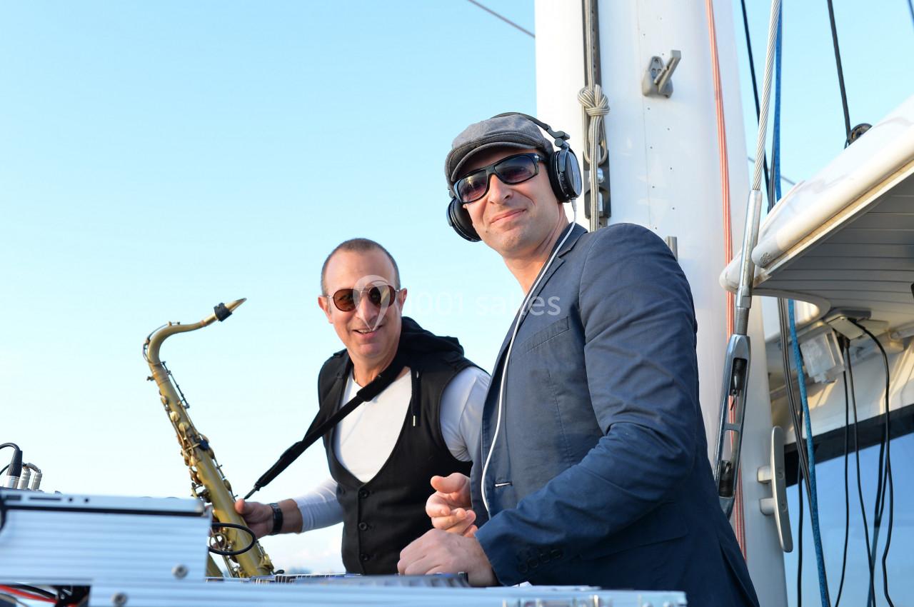 Deux musiciens sur un bateau, l'un jouant du saxophone et l'autre mixant avec un casque audio.