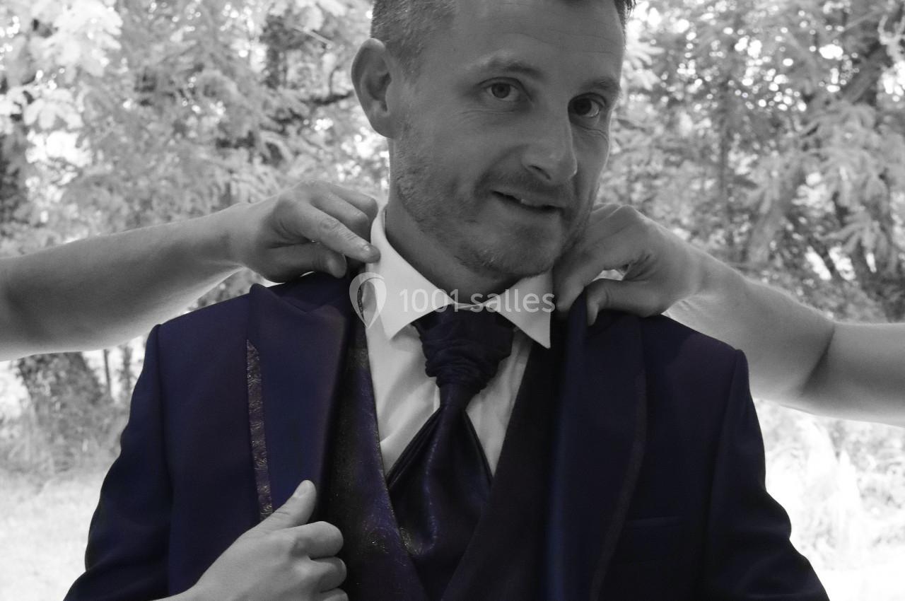 Un homme en costume sombre est aidé par deux personnes ajustant sa veste, devant un fond naturel flou.