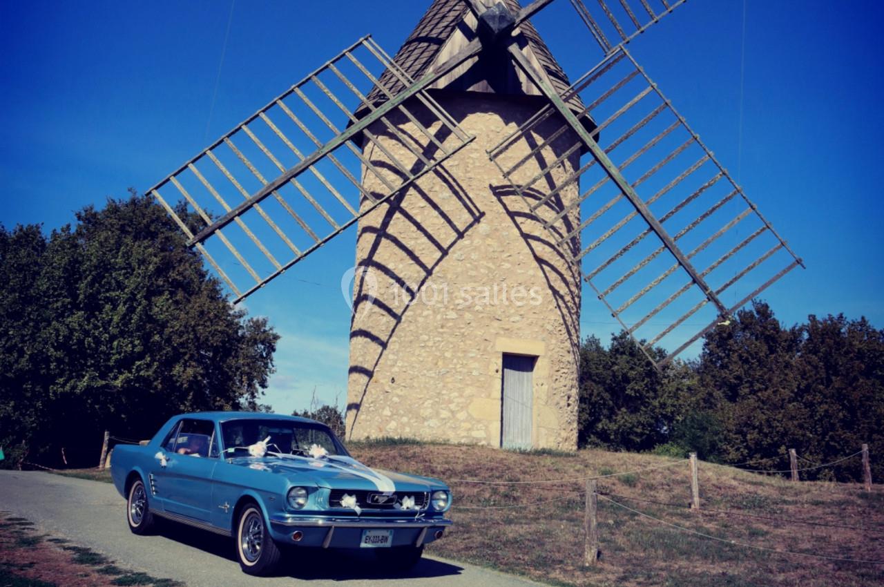 Voiture ancienne bleue garée devant un moulin en pierre avec des ailes en bois, entouré de végétation.