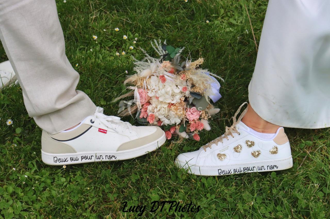 Deux paires de chaussures blanches face à face sur de l'herbe, avec un bouquet de fleurs posé au centre.