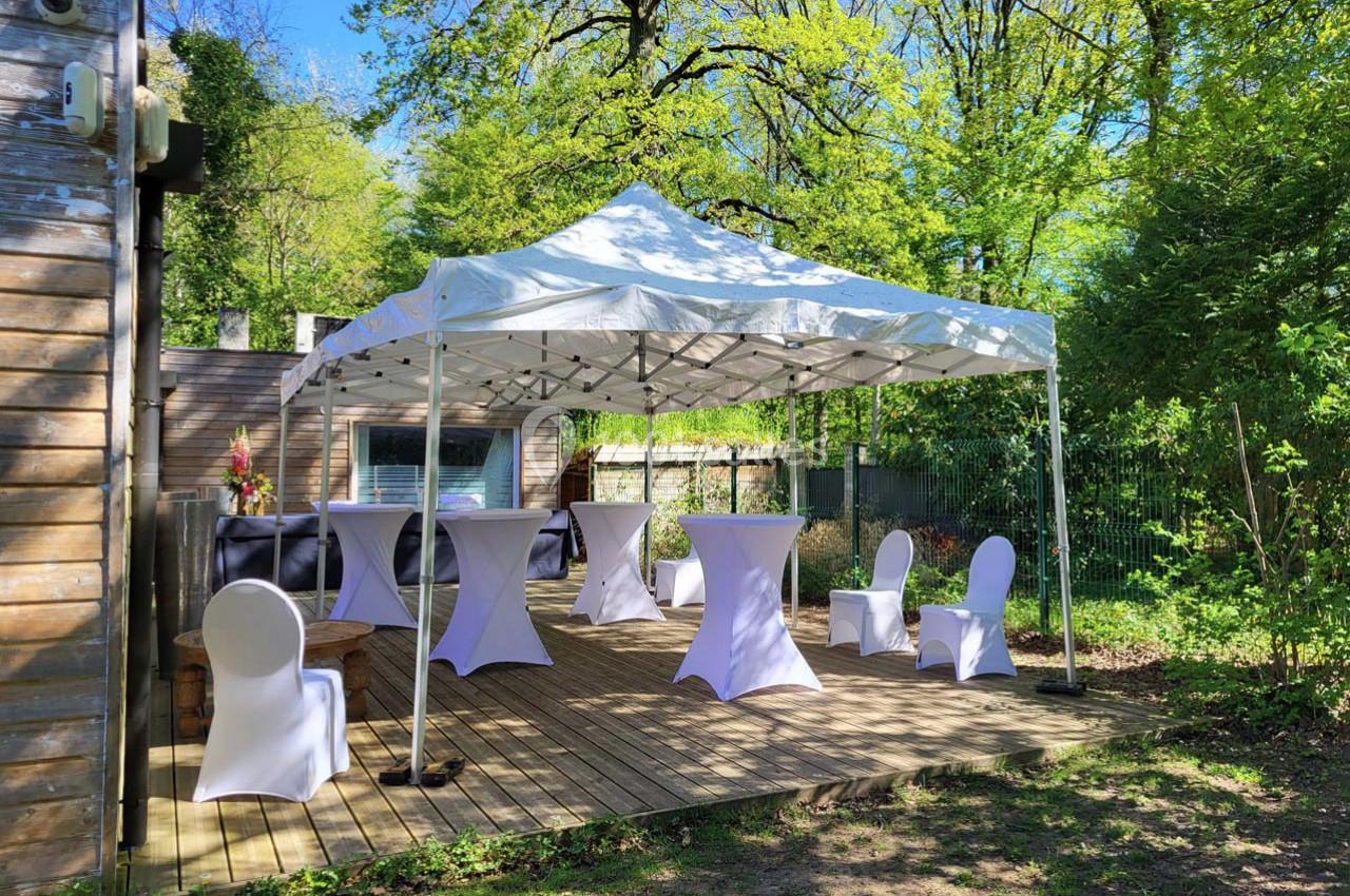 Tente blanche installée sur une terrasse en bois, entourée de verdure, avec tables et chaises habillées de housses.