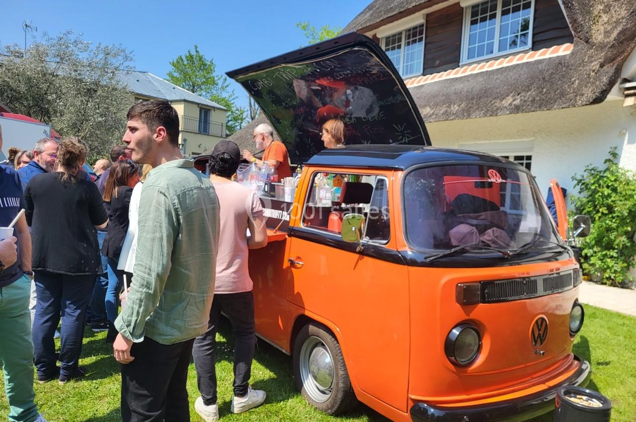 Un van orange aménagé en stand de vente, entouré de personnes dans un jardin par une journée ensoleillée.