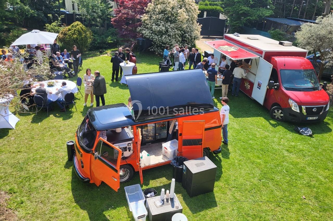 Vue aérienne d'un rassemblement en plein air avec des food trucks et des convives autour de tables sur une pelouse.