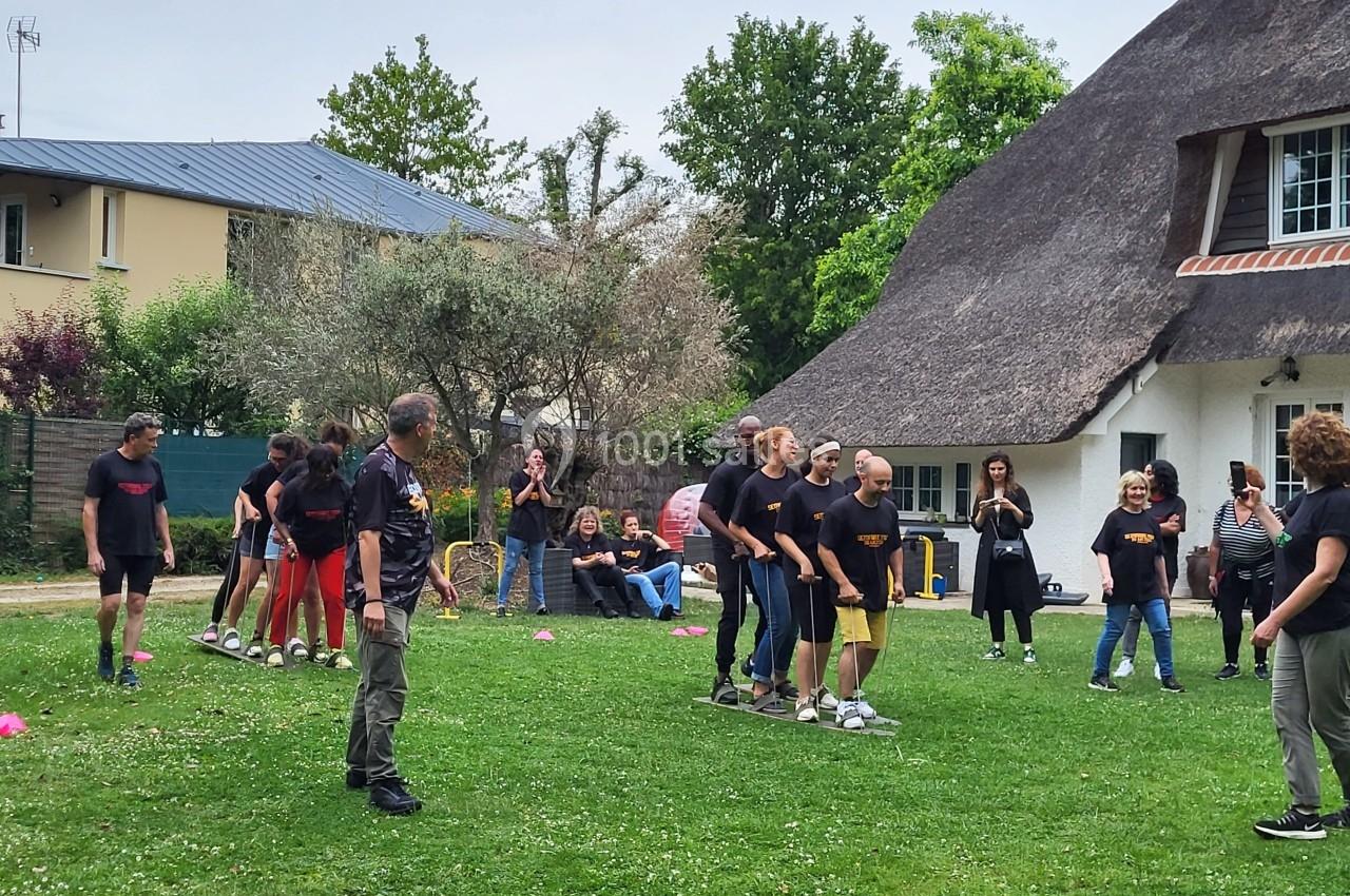 Des personnes participent à un jeu d'équipe en extérieur sur une pelouse, devant des spectateurs et une maison au toit de…