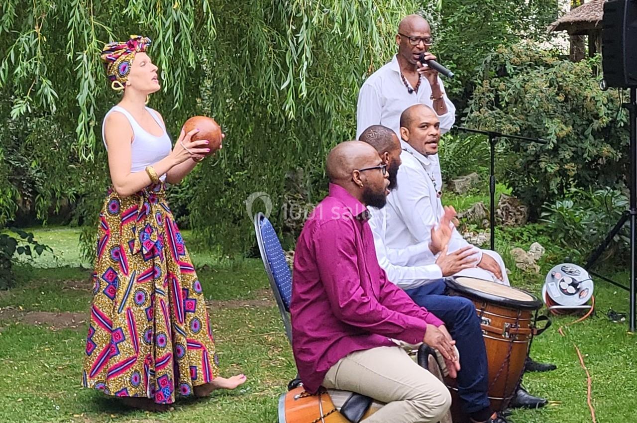 Un groupe de musiciens joue des percussions en plein air, entouré de verdure et d'arbres.