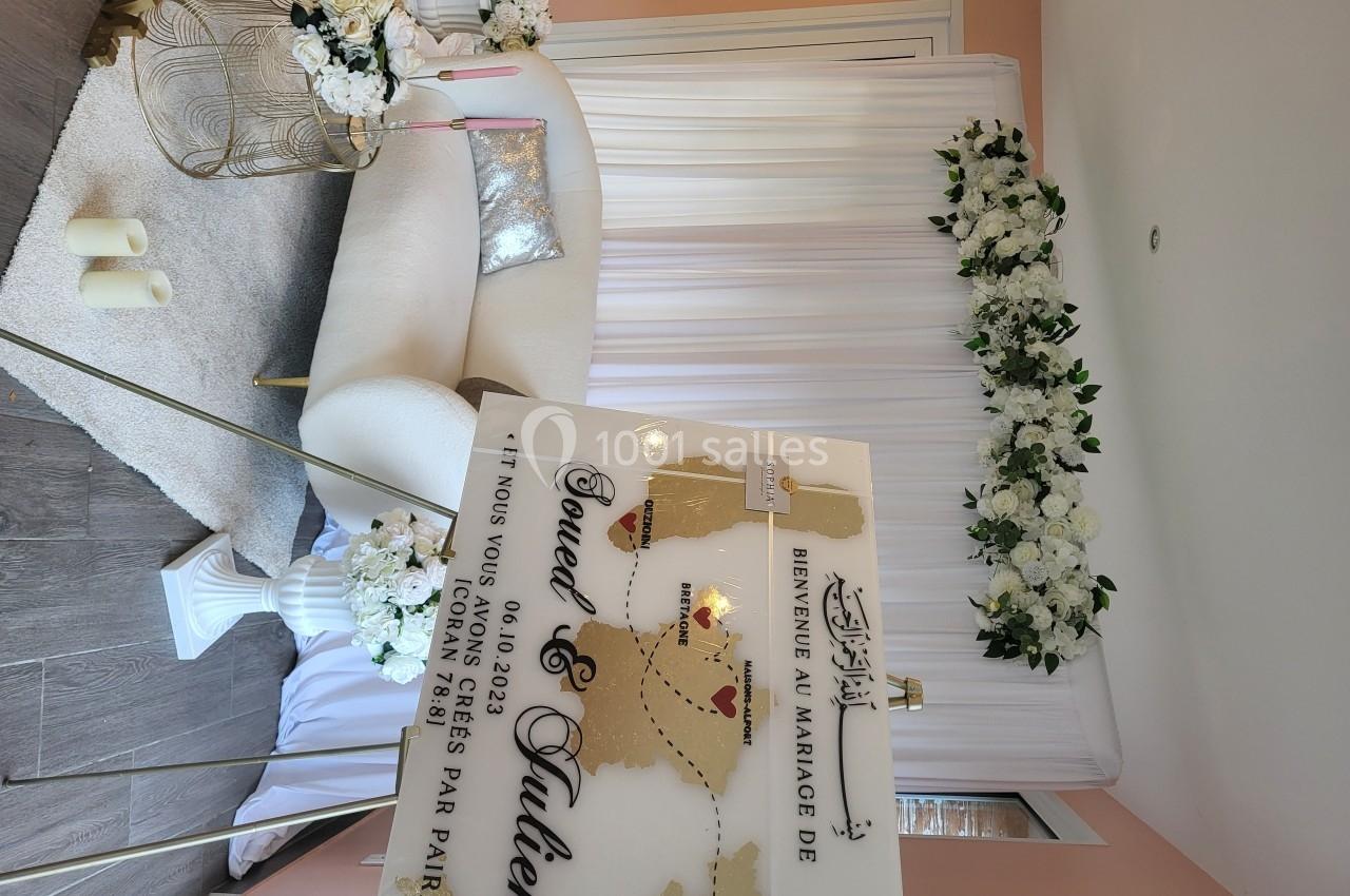 Panneau de bienvenue à un mariage devant un décor floral blanc avec canapé et bougies dans une salle lumineuse.