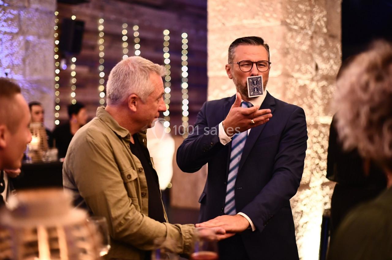 Un magicien en costume réalise un tour de cartes devant un homme dans un cadre festif et lumineux.
