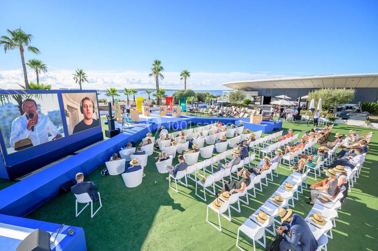 Conférence en plein air avec un public assis, deux écrans géants affichant des intervenants, sous un ciel dégagé.