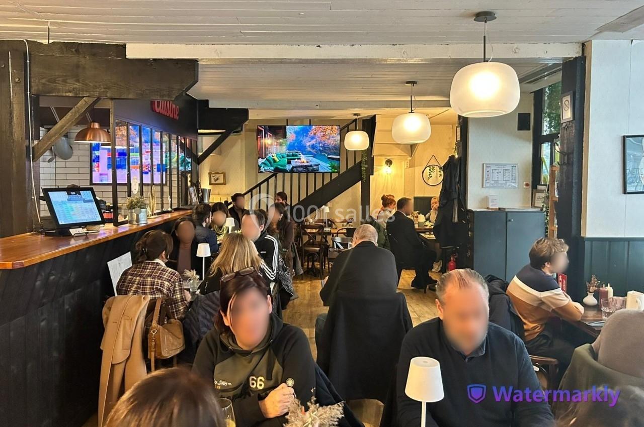 Intérieur d'un café animé avec des clients attablés, un comptoir à gauche et un écran diffusant une image au fond.