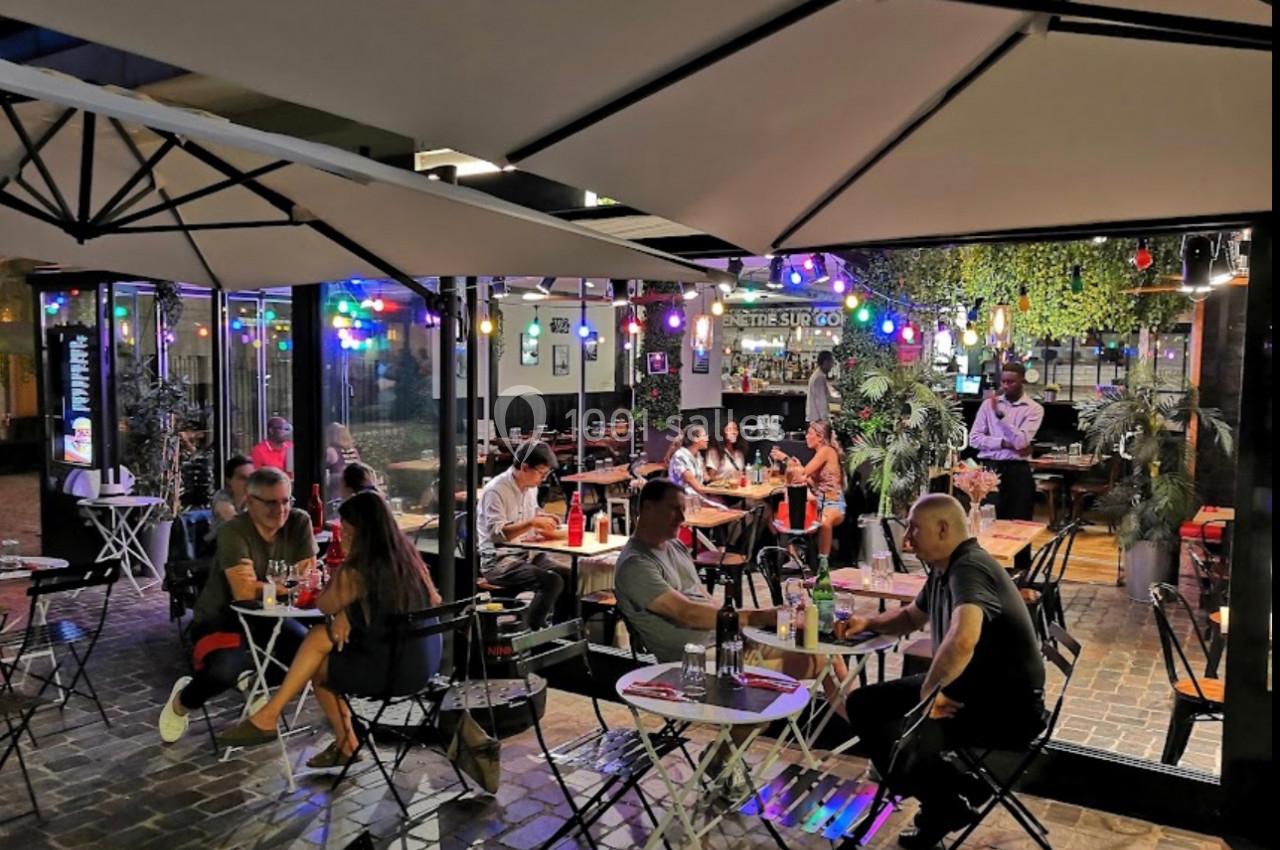 Terrasse d'un restaurant en soirée, avec des clients attablés sous des parasols et des guirlandes lumineuses colorées.