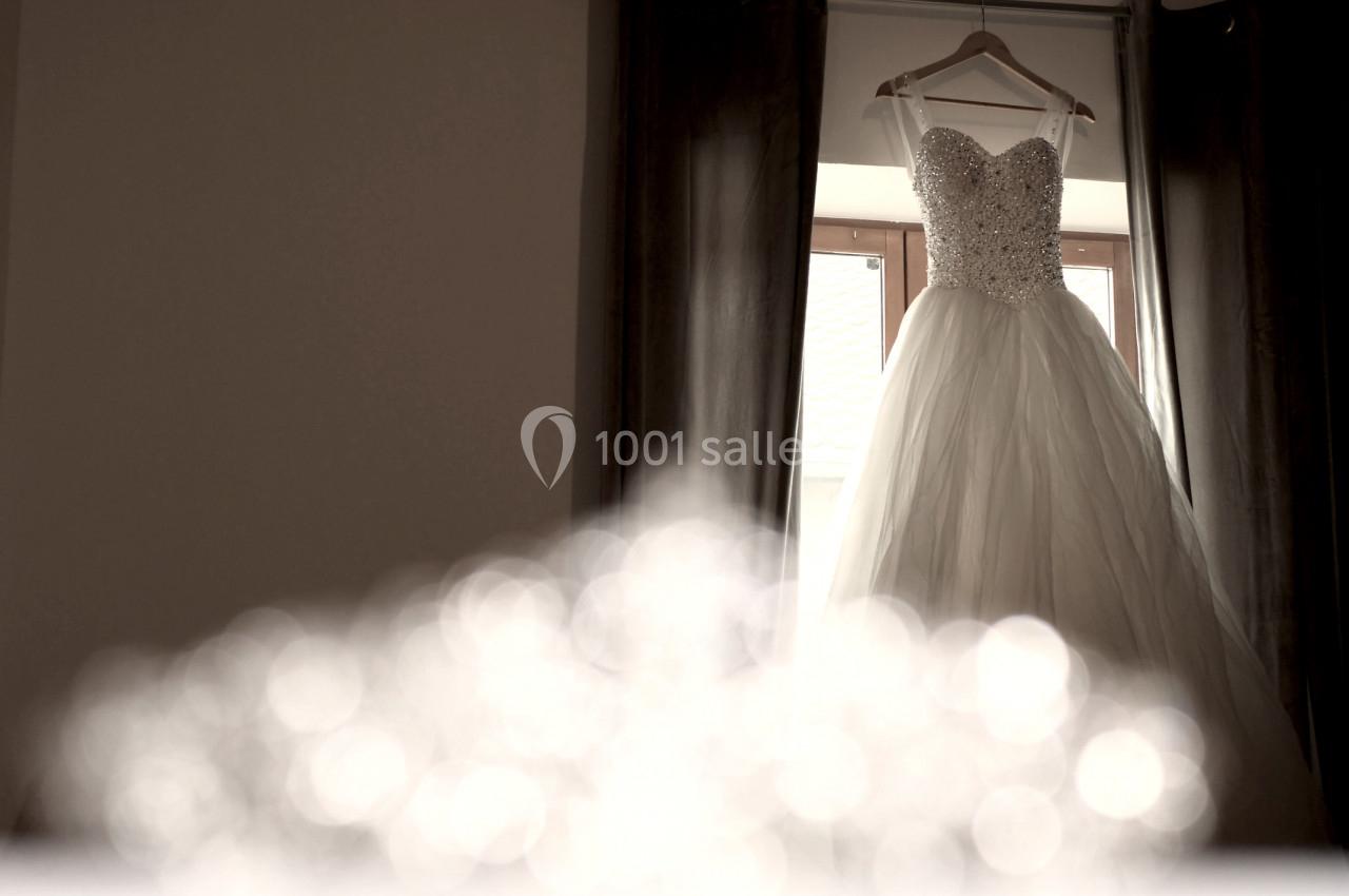 Robe de mariée blanche ornée de perles suspendue à un cintre devant une fenêtre avec des rideaux sombres.
