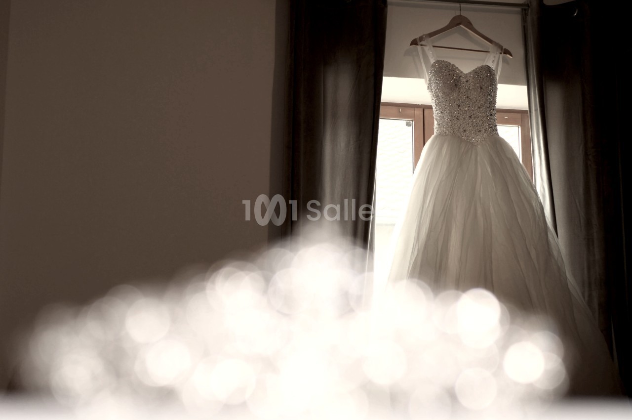 Robe de mariée blanche ornée de perles suspendue à un cintre devant une fenêtre avec des rideaux sombres.