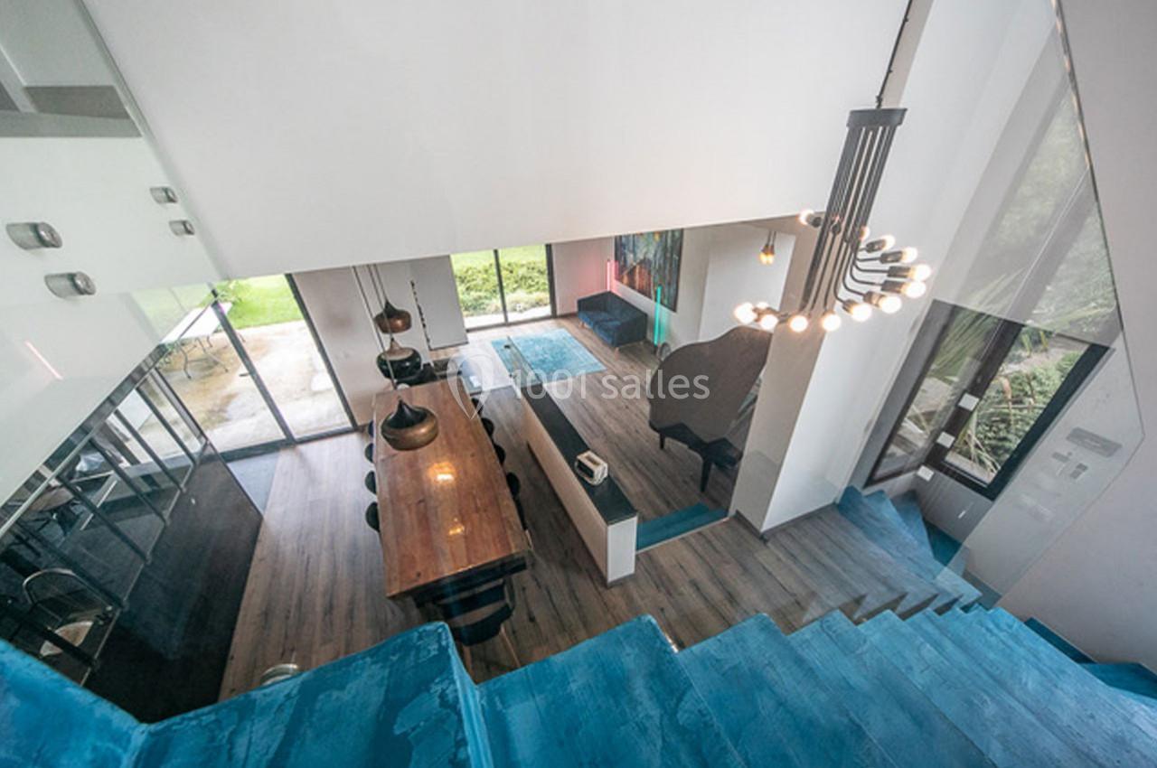 Vue plongeante sur un intérieur moderne avec salle à manger, cuisine ouverte et escalier bleu, donnant sur un jardin.