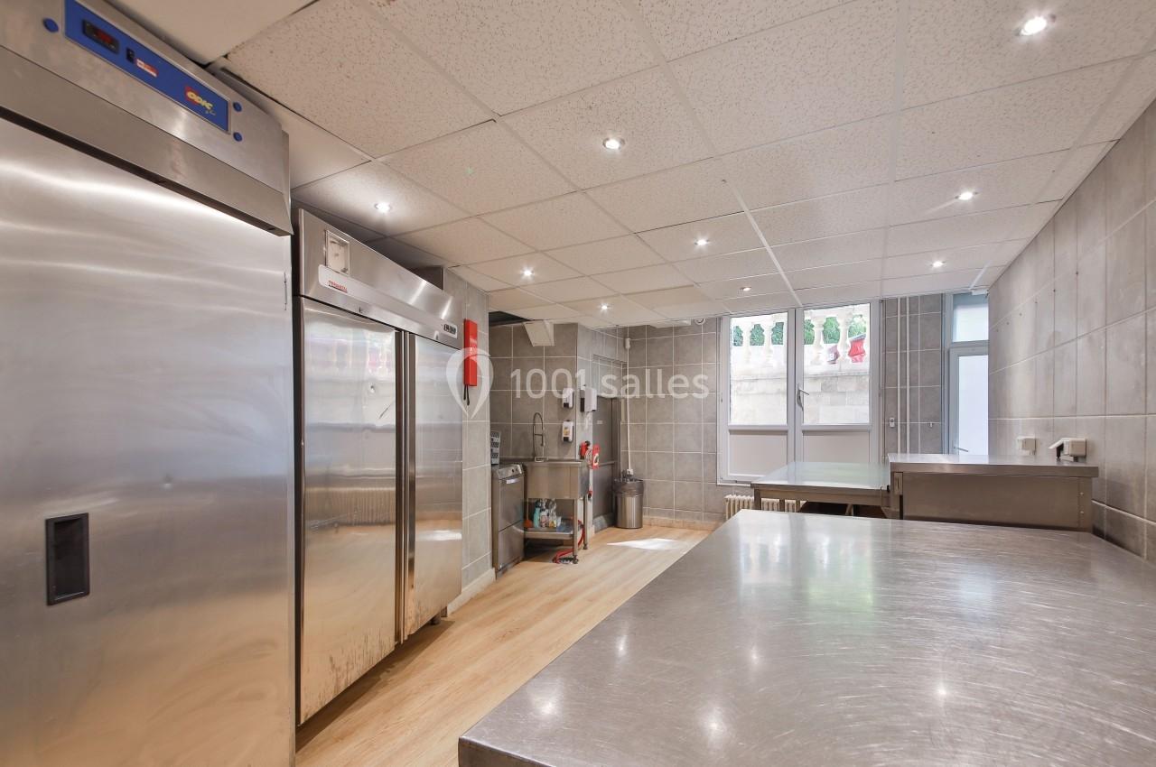 Cuisine professionnelle avec plans de travail en inox, réfrigérateurs, sol en bois clair et murs carrelés gris.