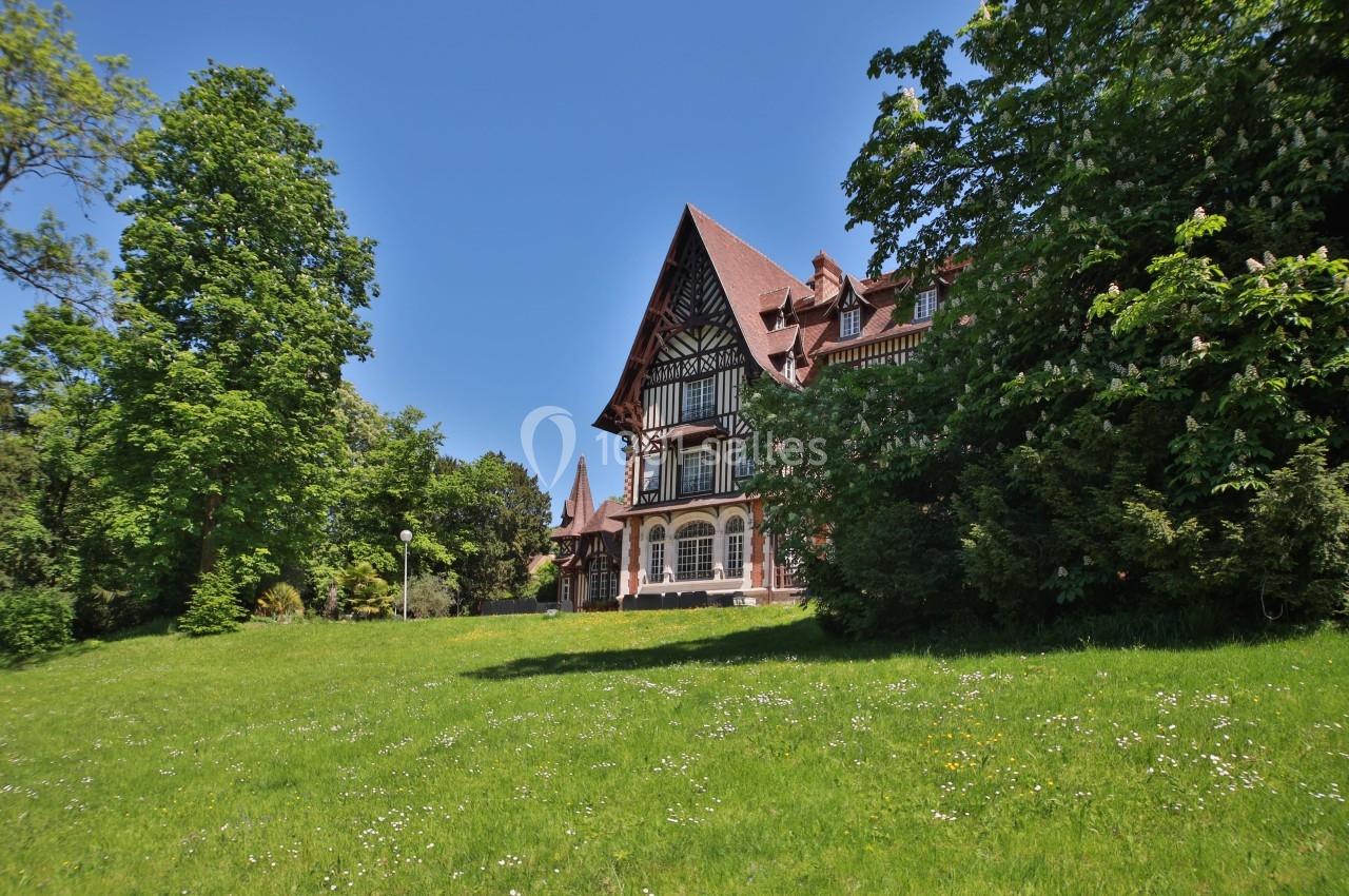 Grande maison à colombages entourée d'arbres et d'une pelouse verdoyante sous un ciel bleu clair.