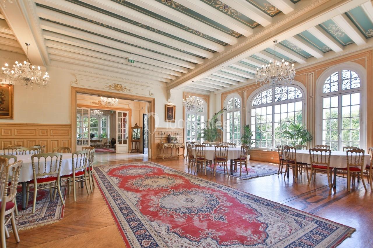 Salle lumineuse avec de grandes fenêtres, tables dressées, chaises élégantes et tapis rouge sur un parquet en bois.