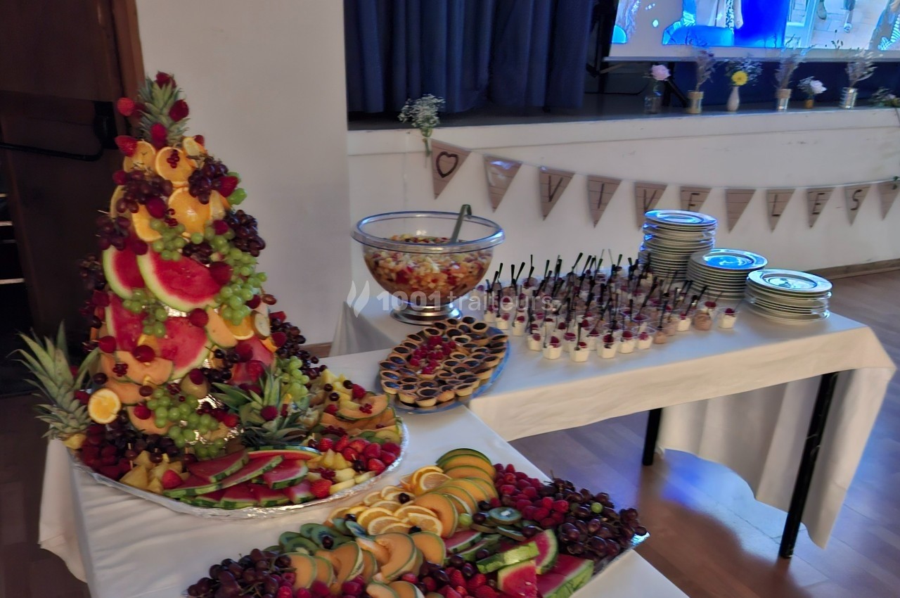 Buffet avec une pyramide de fruits frais, verrines, tartelettes et salade de fruits sur des tables décorées.