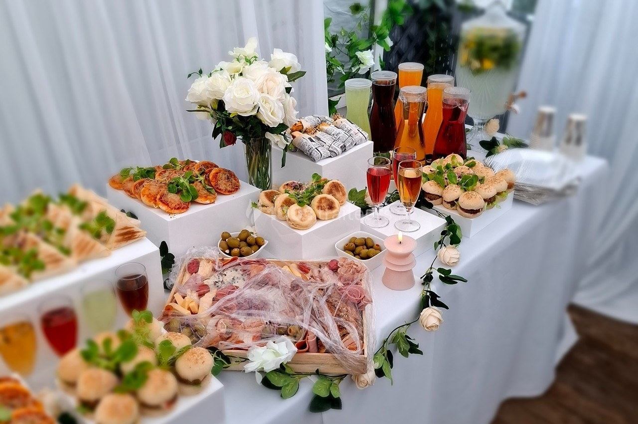 Buffet élégant avec assortiments de charcuterie, pains, olives, boissons variées et décor floral blanc.