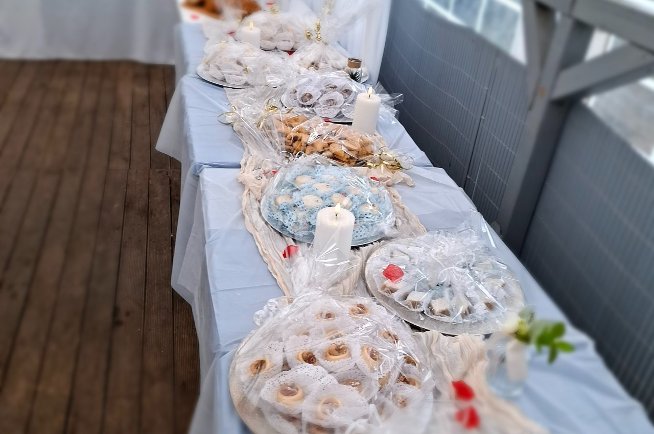 Table garnie de pâtisseries emballées, décorée de bougies blanches et de nappes claires sur une terrasse en bois.