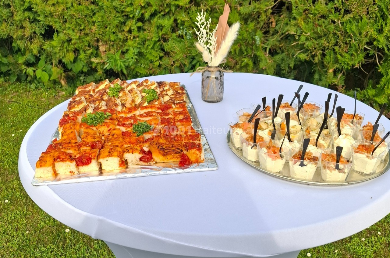 Plateau de bouchées salées variées et verrines apéritives disposés sur une table ronde blanche en extérieur.