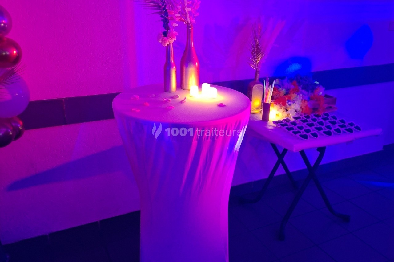 Table haute décorée avec des bougies et des fleurs sous un éclairage violet, à côté d'une table avec des accessoires.