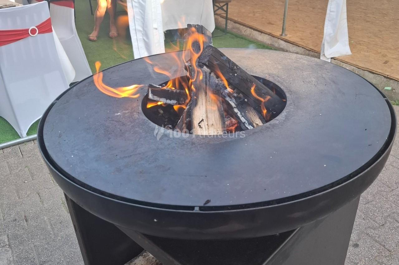Brasero circulaire en métal avec des flammes et des bûches en combustion, installé en extérieur sur un sol pavé.