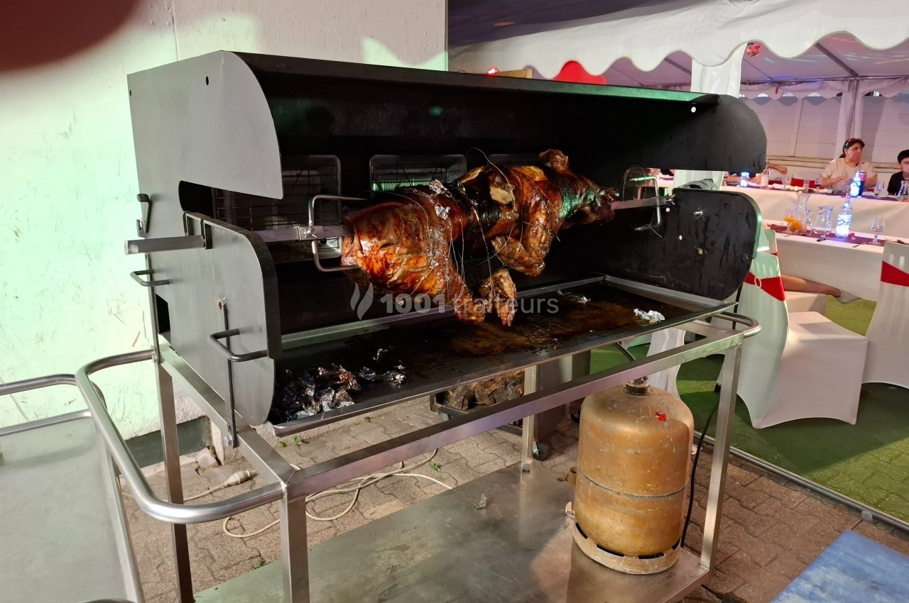 Un cochon entier rôti à la broche dans un barbecue rotatif, avec des tables dressées en arrière-plan.