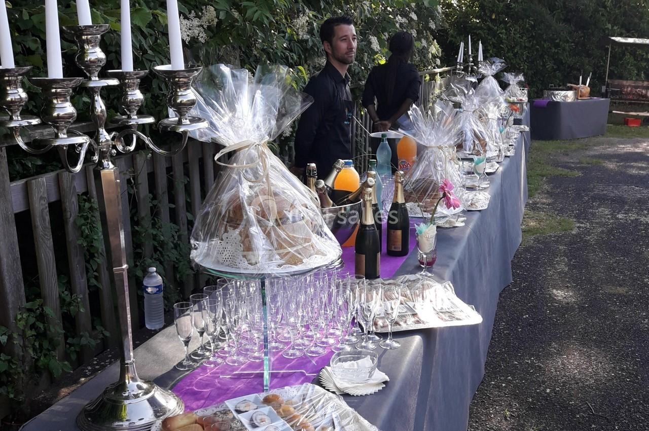 Table de buffet en extérieur avec chandeliers, boissons, viennoiseries et verres, servie par un personnel en tenue noire.