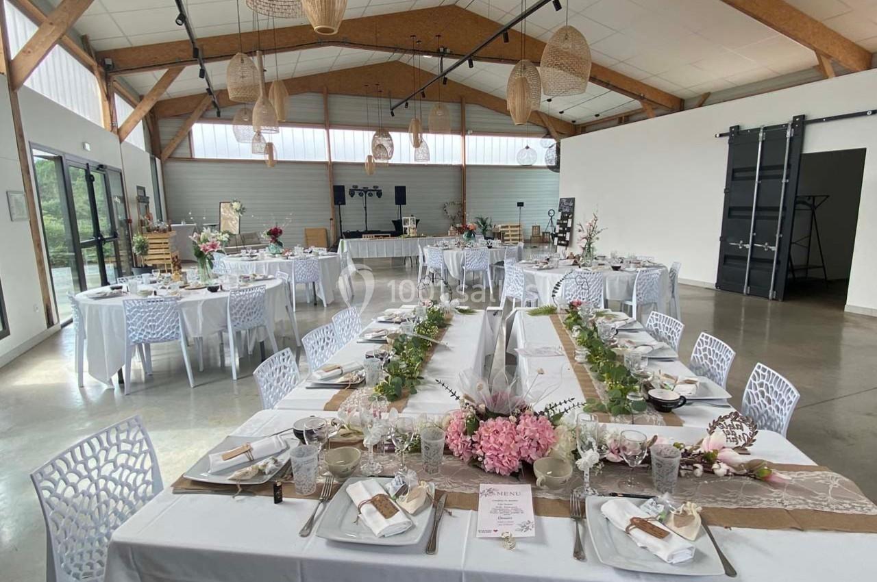 Salle de réception décorée pour un événement, avec tables dressées, fleurs et éclairage naturel.