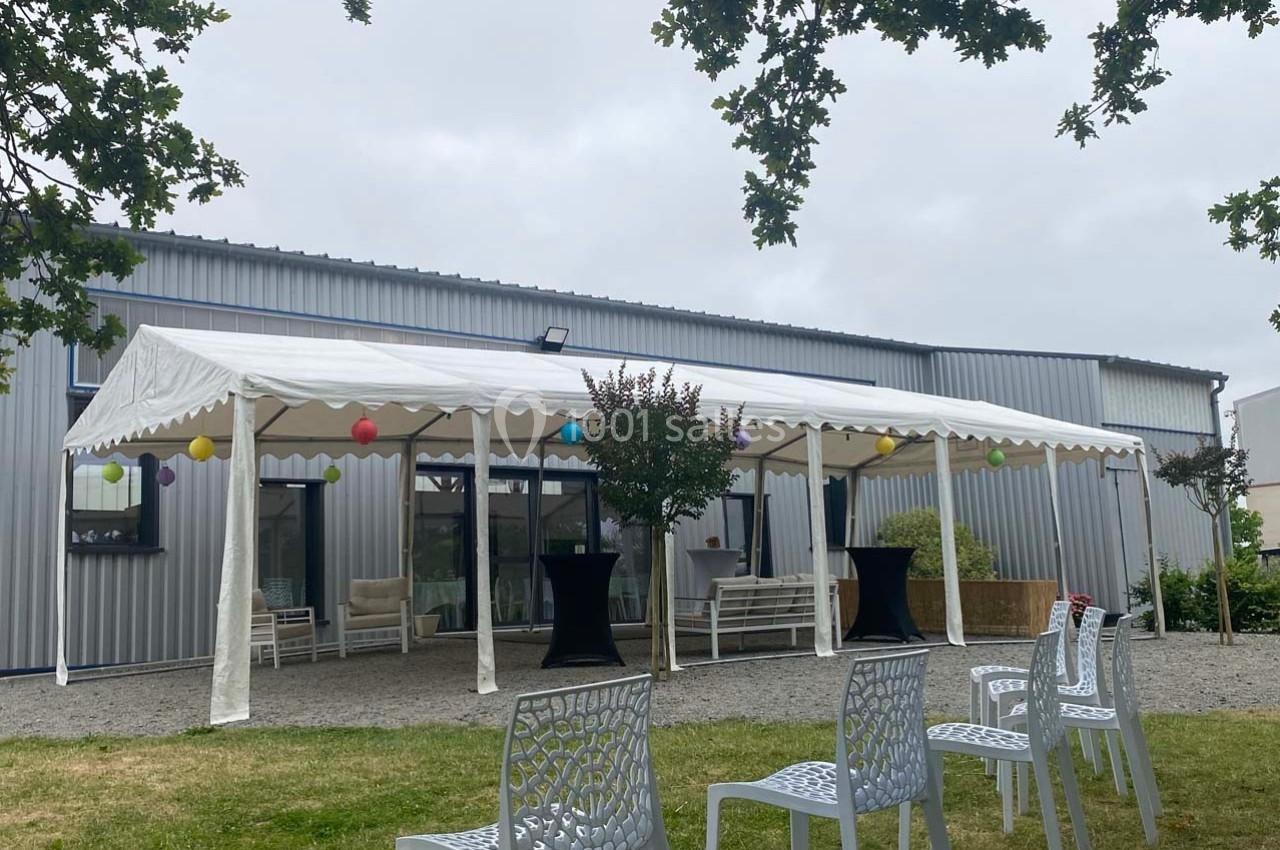 Tente blanche décorée de guirlandes colorées installée devant un bâtiment métallique, avec des chaises sur la pelouse.
